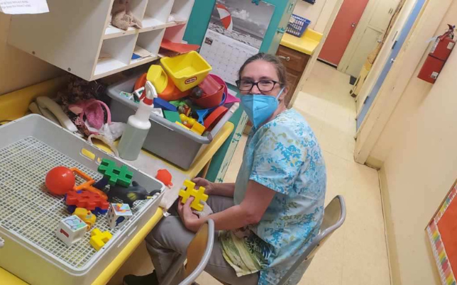 A young woman with her hair pulled back and wearing a mask, glasses and a blue scrub top sits in front of a tub filled with large plastic items